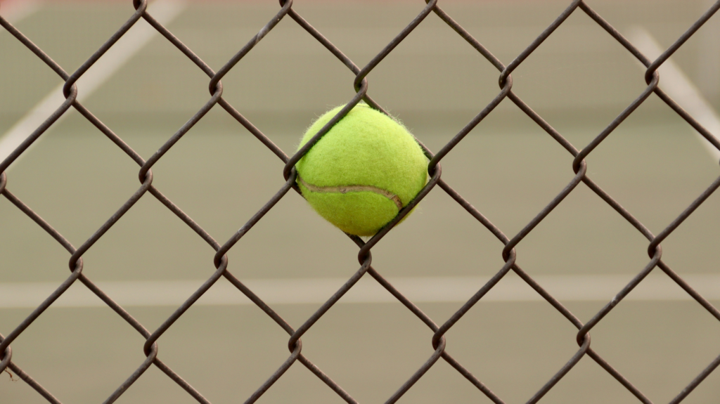 A yellow-green tennis ball wedged tightly in the diamond-shaped opening of a metal chain-link fence near a court.
