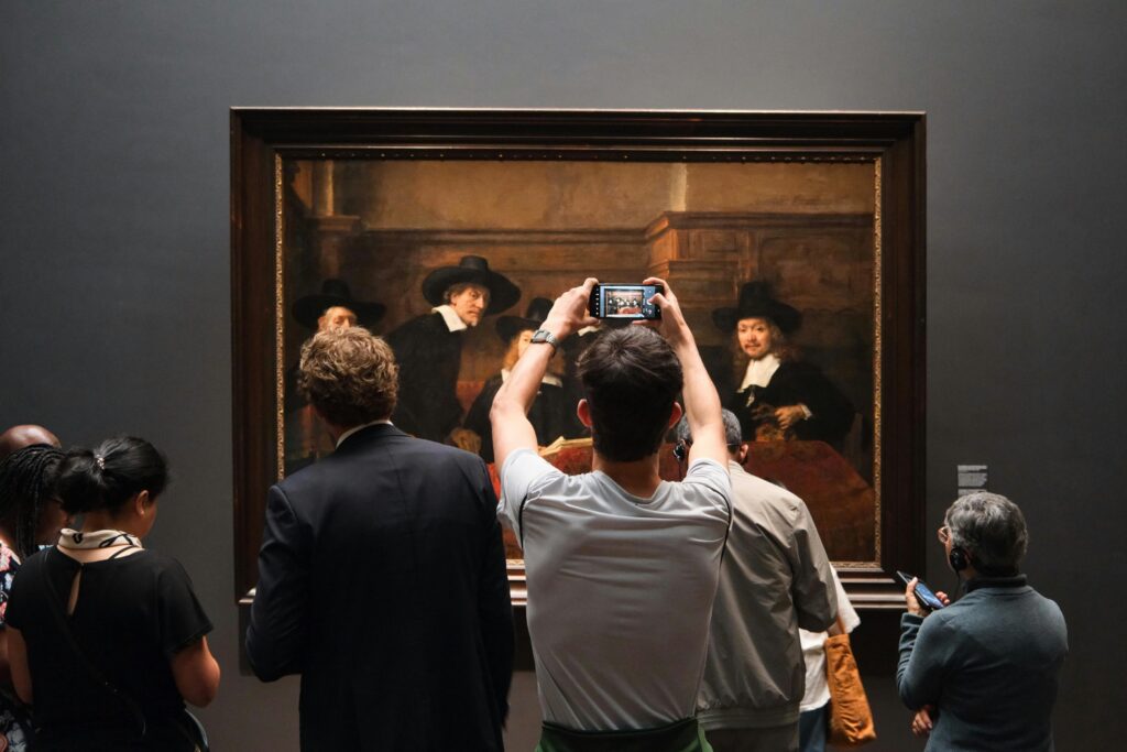 Museum visitors gather before a large Rembrandt painting as one person photographs the artwork with a smartphone.