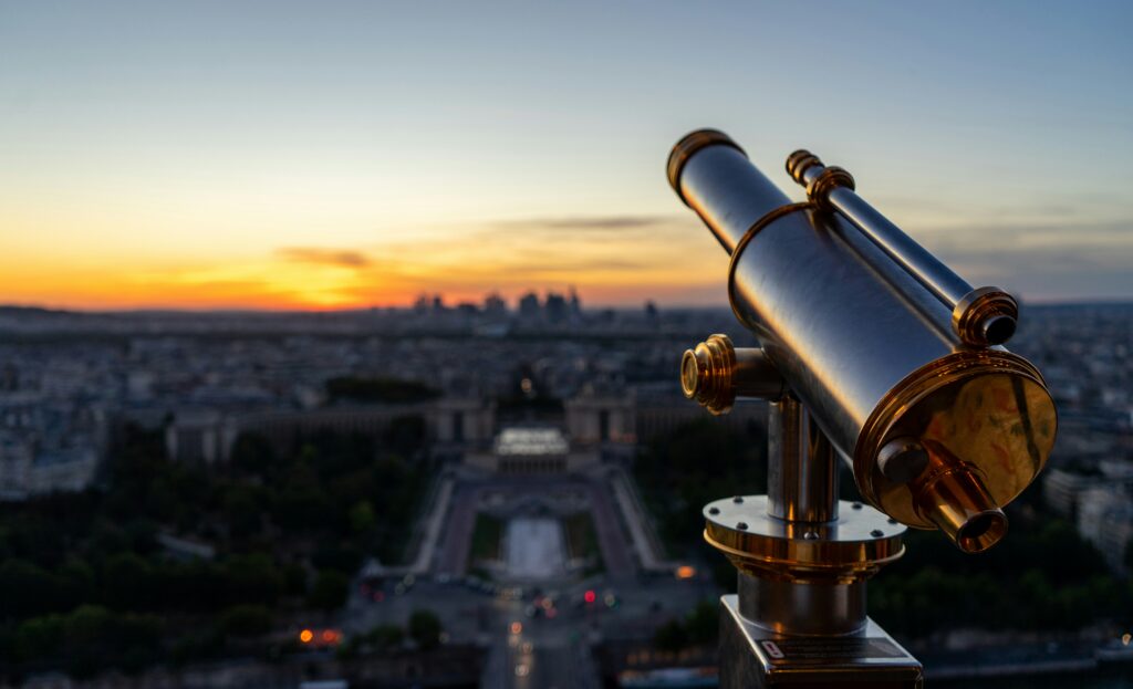 Brass telescope on the Eiffel Tower overlooking the Paris cityscape and Trocadéro gardens at sunset.