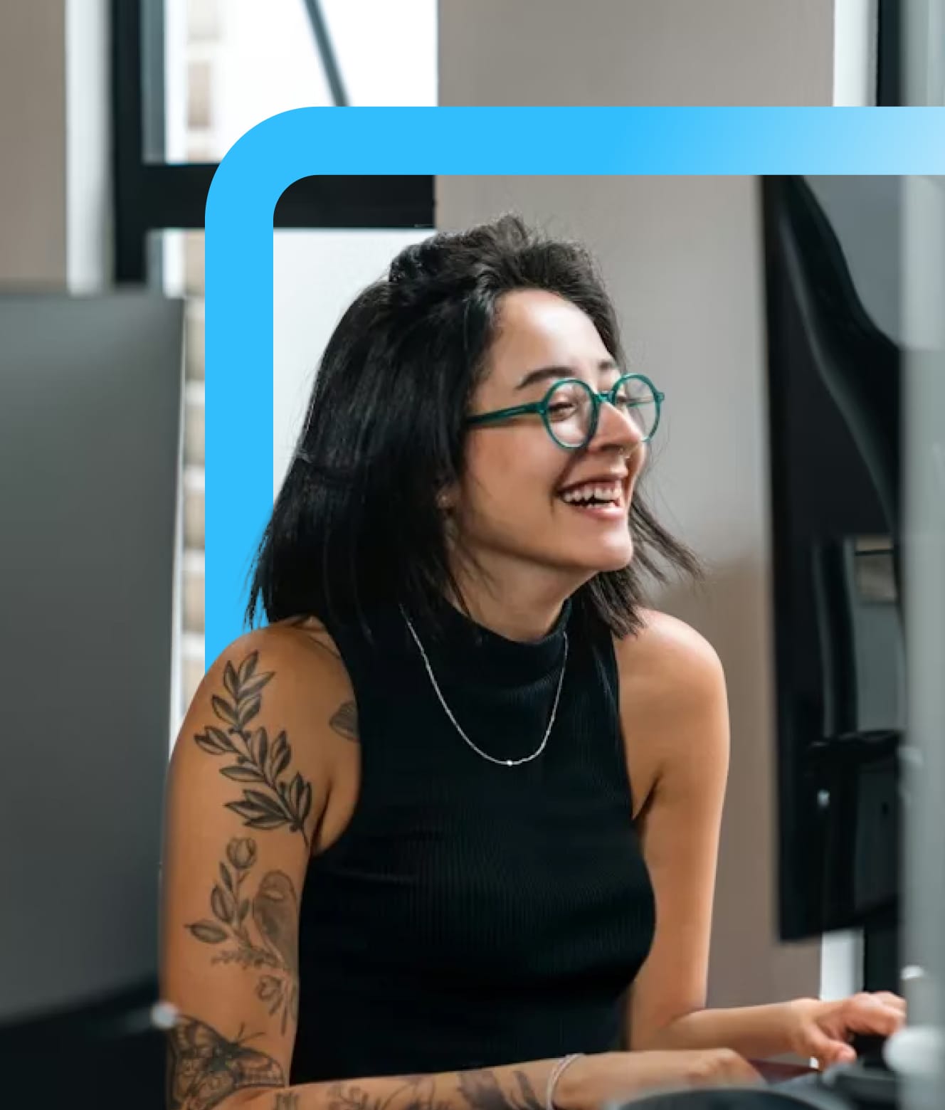 Smiling woman with green glasses and floral arm tattoos working at a desk with computer monitors.