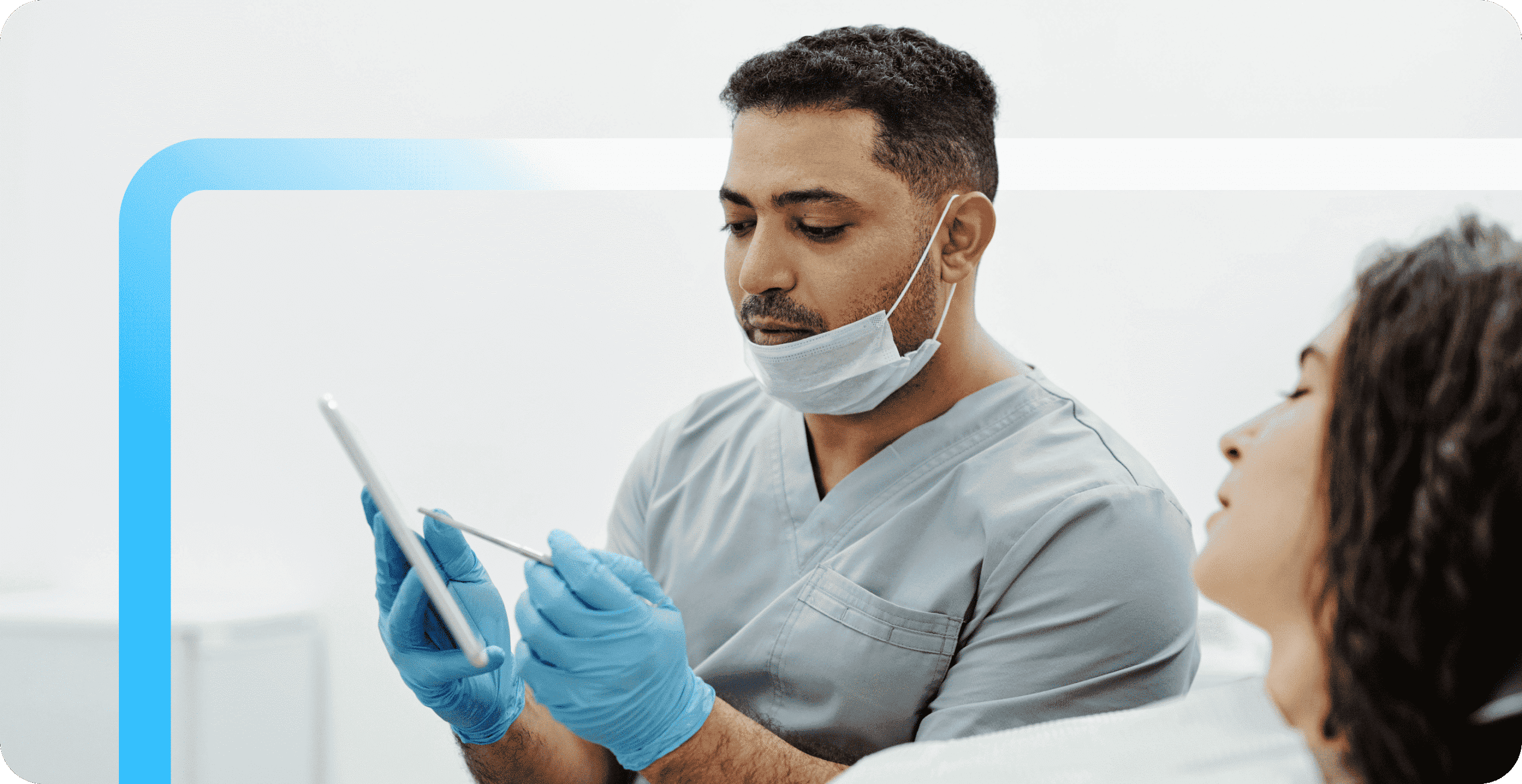 A dental professional in gray scrubs and blue gloves reviews a tablet while consulting with a female patient.