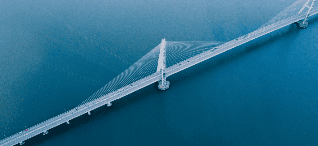 Aerial view of a cable-stayed bridge with a tall central pylon stretching diagonally across calm blue water.